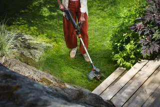 Person trimming lawn edges with a grass trimmer beside a wooden deck and garden plants.