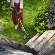 Person trimming lawn edges with a grass trimmer beside a wooden deck and garden plants.