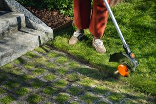 Person trimming a lawn edge with a grass trimmer beside stone steps and grid pavers.