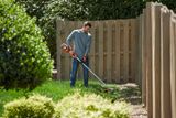 A person using a string trimmer to cut grass along a wooden fence in a garden.