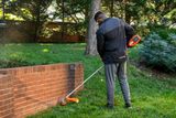 Une personne utilise un coupe-bordure pour border une pelouse le long d’un mur de briques dans un jardin.