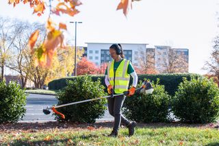 A person using a string trimmer to maintain a landscaped area near a parking lot.