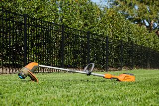 A string trimmer lying on a neatly trimmed lawn with a metal fence and trees in the background.