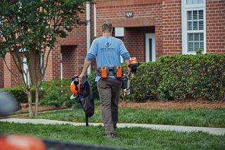 En person, der går på en græssti med haveredskaber med en murstensbygning i baggrunden.