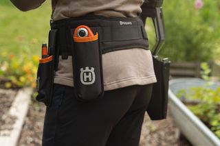 Eine Person, die einen Werkzeuggürtel mit Husqvarna Branding trägt und in einem Garten steht.