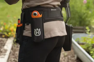 En person som bär ett verktygsbälte med Husqvarna-logotyp och står i en trädgård.
