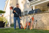 A person holding a garden trimmer, with various gardening tools including a leaf blower and hedge trimmer arranged against a brick wall in a backyard setting.