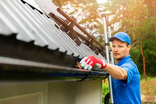 A person cleaning leaves from a house gutter using a ladder.