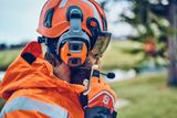 Une personne portant un équipement de travail et un casque de sécurité avec protection d’oreilles et visière, communiquant via un casque dans un environnement extérieur.
