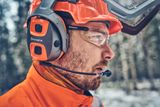 Nahaufnahme einer Person, die einen Husqvarna Helm mit Gehörschutz und Visier in einer Waldumgebung trägt.