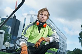 A worker in a green work wear holding a tool, with a building in the background.