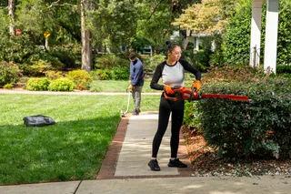 A person trimming bushes with a hedge trimmer, while another person uses a string trimmer on a lawn.