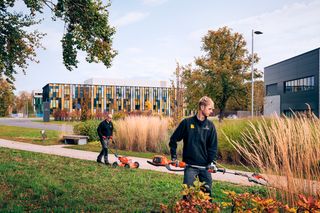 Two people working with outdoor equipment on a landscaped path near modern buildings.