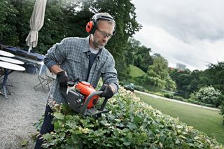 Henkilö, joka leikkaa pensasaitaa pensasleikkurilla puutarhaympäristössä.