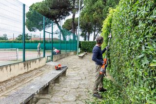 A person trimming a tall hedge with a pole hedge trimmer near a tennis court.