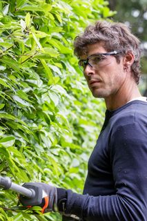 A person trimming a hedge with a long-handled hedge trimmer in a garden.