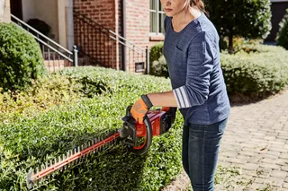 A person trimming a hedge with a hedge trimmer in a residential garden.