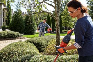 Zwei Personen, die Gartengeräte verwenden, um Büsche zu trimmen und einen Garten zu pflegen.