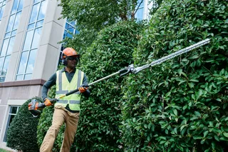 A person using a hedge trimmer to shape bushes in front of a building.