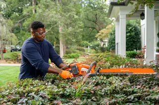 A person trimming hedges with a Husqvarna hedge trimmer in a garden setting.
