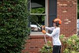 A person using a hedge trimmer to trim bushes near a brick house.