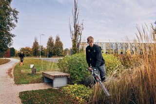 Uma pessoa a utilizar um corta-sebes num ambiente de parque, com outra pessoa a cortar a relva ao fundo.