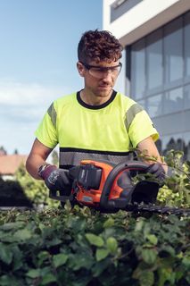 A person using a hedge trimmer to trim bushes outside a modern building.