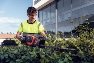 Une personne qui utilise un taille-haie pour tailler des buissons à l’extérieur d’un bâtiment moderne.
