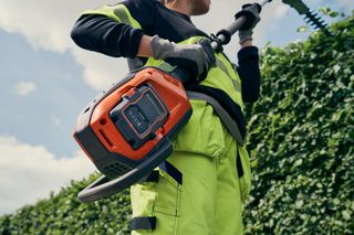 Eine Person, die eine Heckenschere verwendet, um eine hohe Hecke zu schneiden, wobei der Schwerpunkt auf dem Batteriefach liegt.