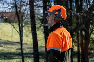 Person iført Husqvarna-arbejdshjelm med høreværn og ansigtsværn i skovomgivelser.