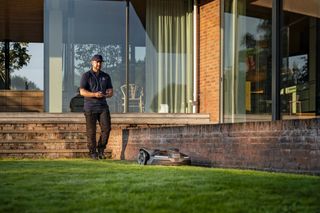 Eine Person, die in einem Garten steht und einen Mähroboter in der Nähe eines modernen Hauses überwacht.