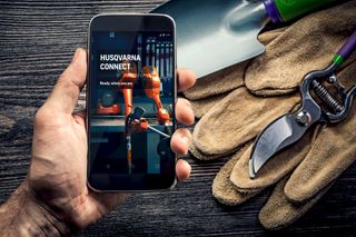 A hand holding a smartphone displaying the Husqvarna Connect app, with gardening tools in the background.