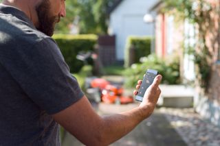 A person using a smartphone to control a robotic lawn mower in a garden setting.