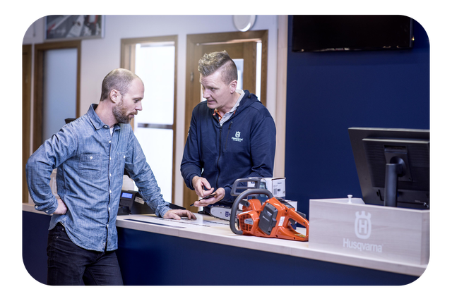 Two people at a counter discussing a Husqvarna chainsaw, with a display monitor and Husqvarna signage visible.