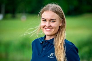 Asmuo, vilkintis "Husqvarna" polo marškinėlius, stovintis lauke, ant vejos.