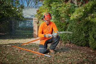 A person in work gear assembling a pole saw in a garden setting.