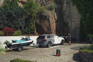 Husqvarna PW 360. Une personne lave une voiture avec un nettoyeur haute pression dans un environnement extérieur rocheux, avec un jet ski sur une remorque à proximité.