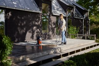 Husqvarna PW 130. Une personne utilise un nettoyeur haute pression pour nettoyer une terrasse en bois à l’extérieur d’une maison.