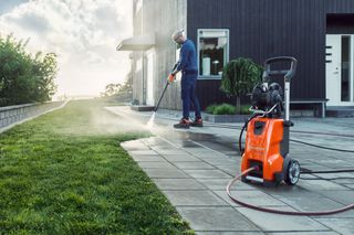 A person using a pressure washer to clean a patio next to a modern house.