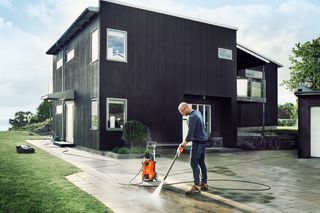 En person som använder en högtryckstvätt för att rengöra en asfalterad uppfart framför ett modernt hus.
