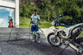 A person cleaning a motorcycle with a pressure washer in a garden setting.