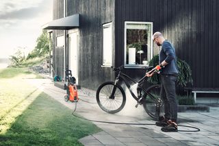 A person using a pressure washer to clean a bicycle on a patio.