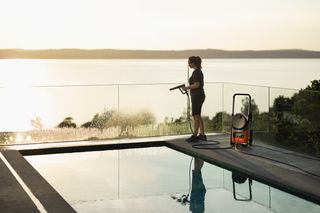Husqvarna PW 370. Une personne utilisant un nettoyeur haute pression pour nettoyer une clôture en verre au bord d’une piscine au coucher du soleil.
