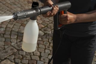 A person using a pressure washer with a detergent bottle attached, spraying water on a cobblestone surface.