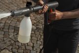 Een persoon die een hogedrukreiniger gebruikt met een wasmiddelfles eraan, water spuit op een geplaveide ondergrond.