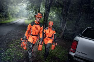 Two people in work gear carrying chainsaws and fuel cans near a forest road.