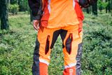 Людина в штанях Husqvarna для роботи з ланцюговою пилкою в лісі, що стоїть обличчям убік.