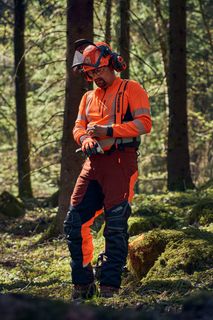 A person in work gear standing in a forest, holding a chainsaw.