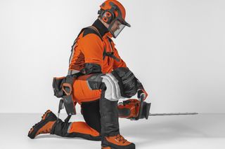 Sidovy av en person i arbetsutrustning som knäböjer med en motorsåg, som visar skyddande knäskydd.