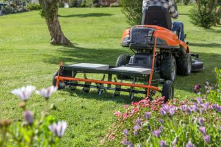 Eine Person, die einen Aufsitzmäher mit einem Aerifizierer-Anbaugerät in einem Garten verwendet.